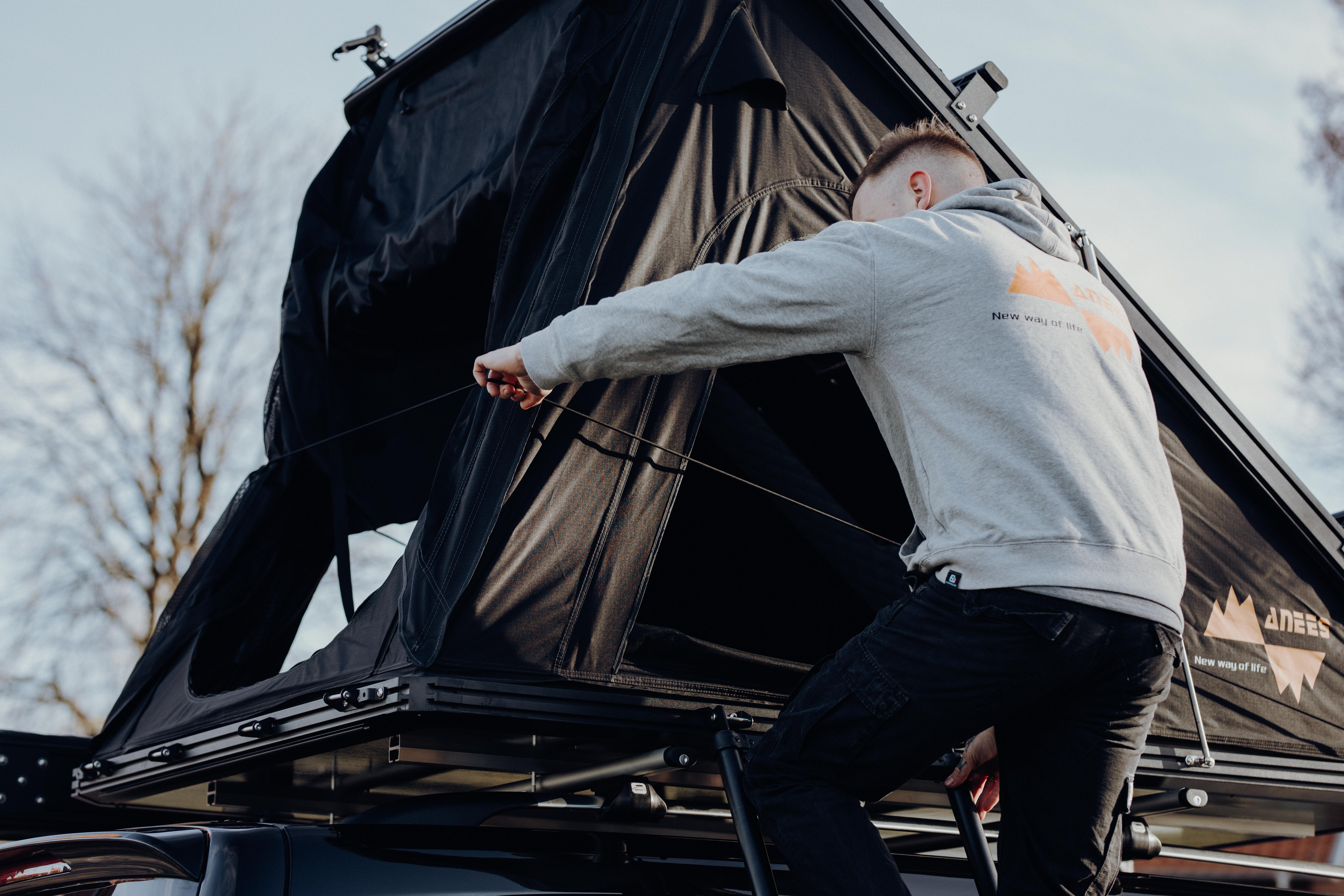 Mann im grauen Hoodie auf Leiter befestigt den Stoff eines schwarzen Dachzelts am Fahrzeug