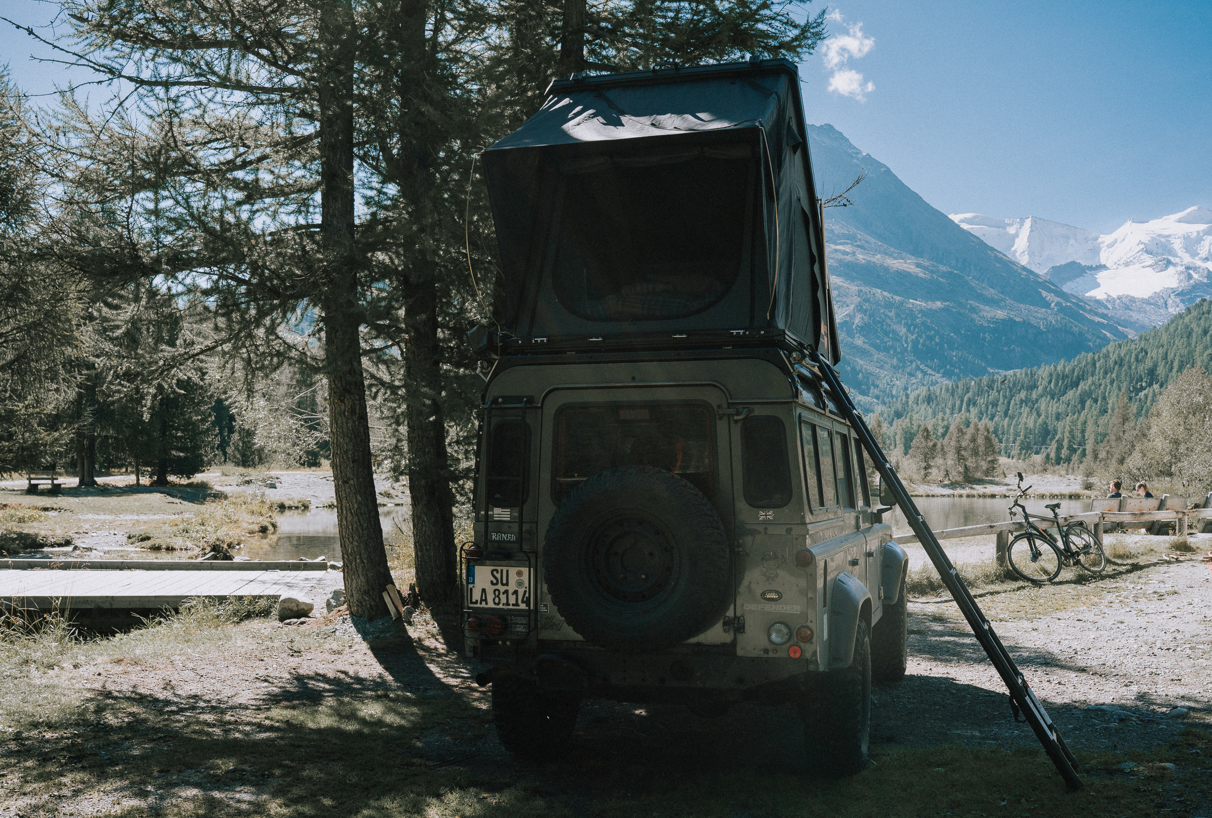 Camper mit Dachzelt am kleinen See zwischen Kiefern, daneben ein Fahrrad, im Hintergrund schneebedeckte Berge