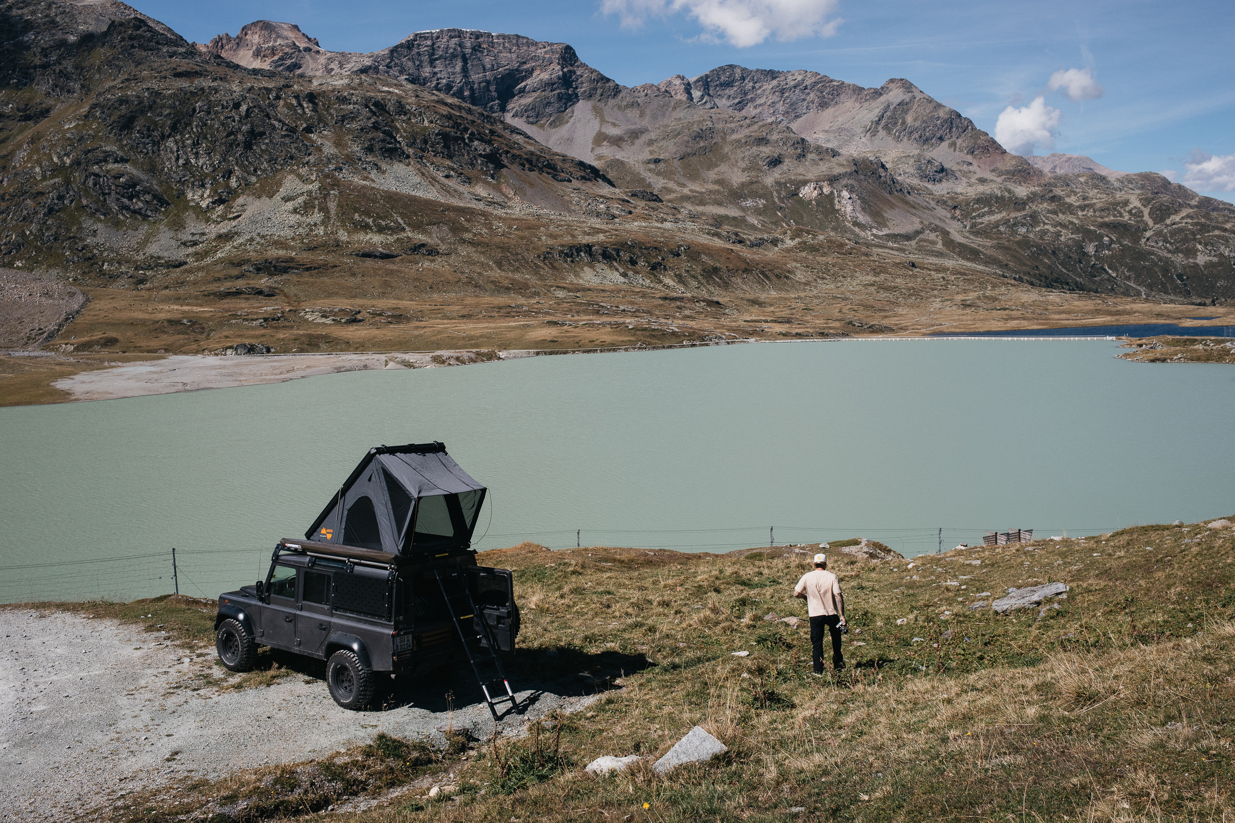 Geländewagen mit Dachzelt am alpinen See, eine Person geht am grasigen Ufer vor schroffen Bergen entlang