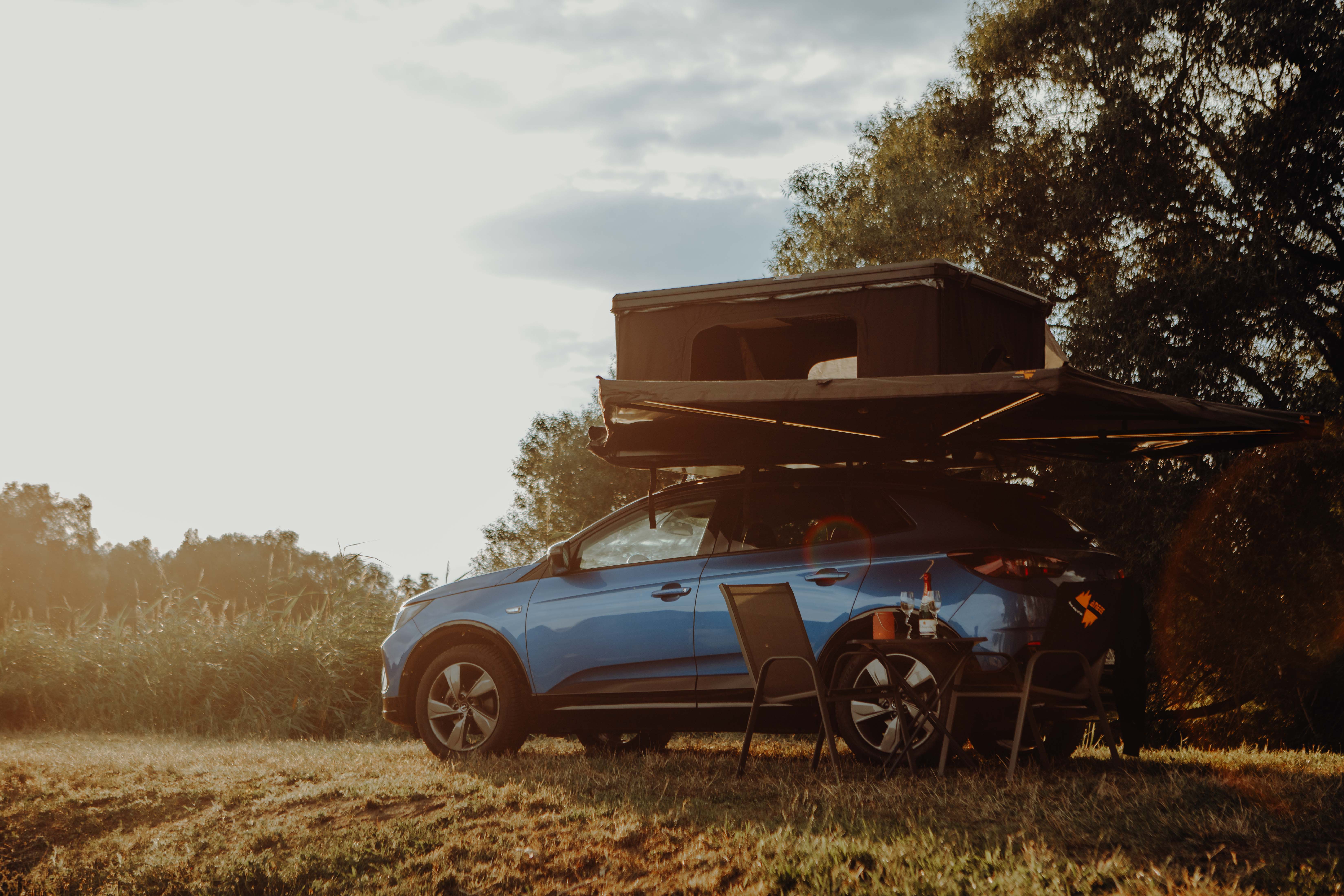 Blauer SUV mit Dachzelt und Markise steht bei Sonnenuntergang auf einer Wiese neben Tisch und Stühlen.