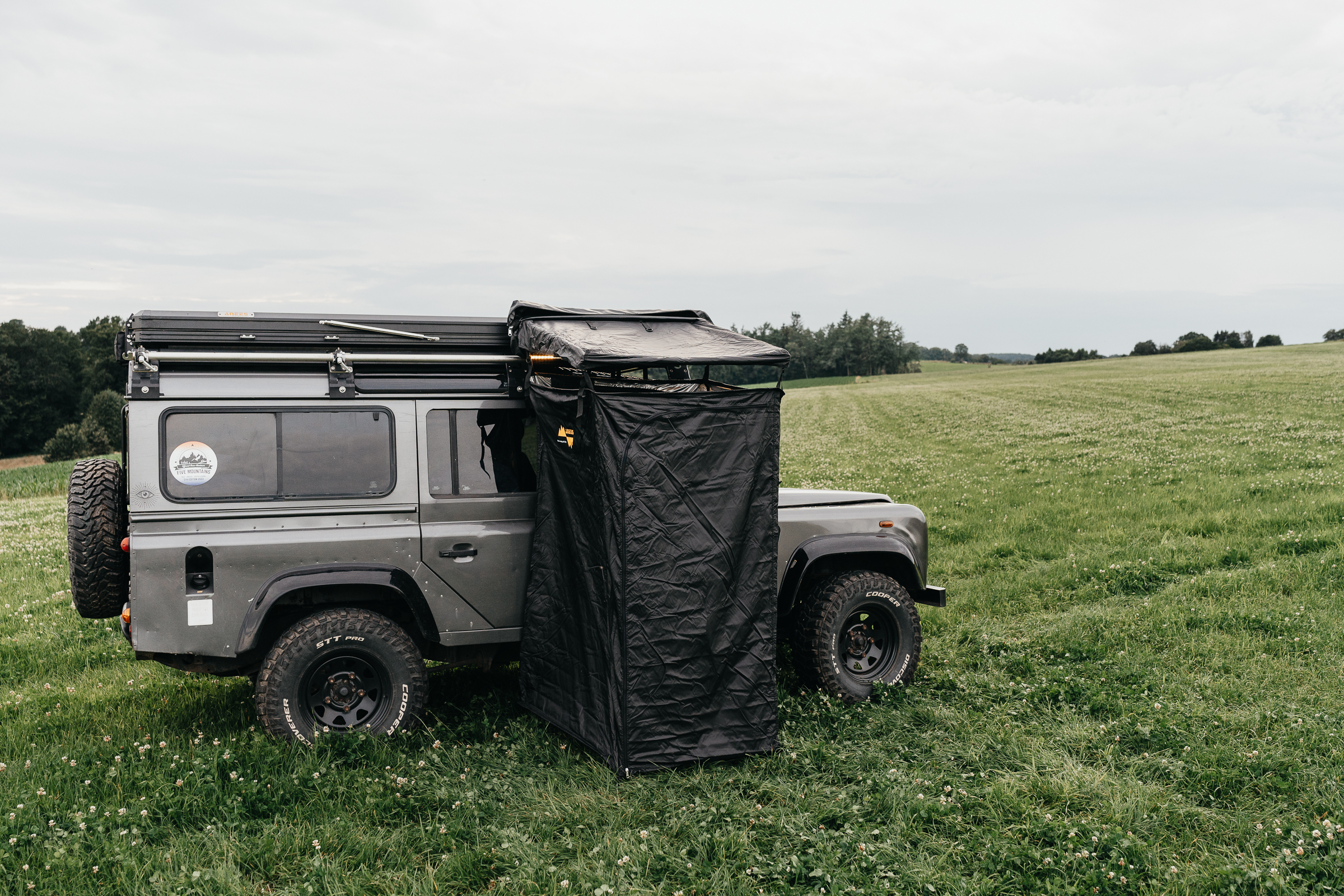 Grauer Geländewagen mit Dachzelt und schwarzem Sichtschutzzelt auf einer Wiese unter bewölktem Himmel